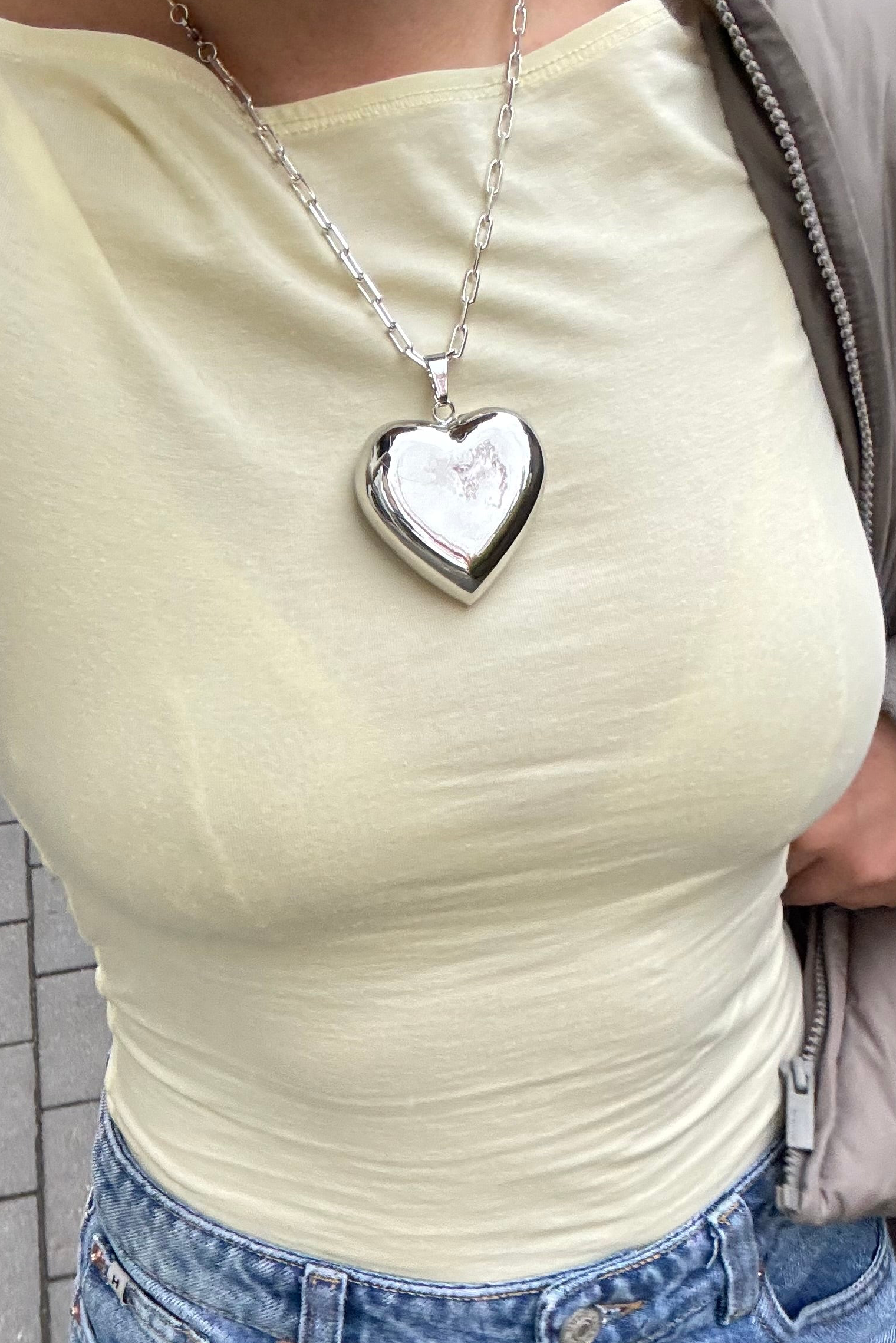 Heart-shaped silver pendant on a necklace worn by a person in a light yellow top.