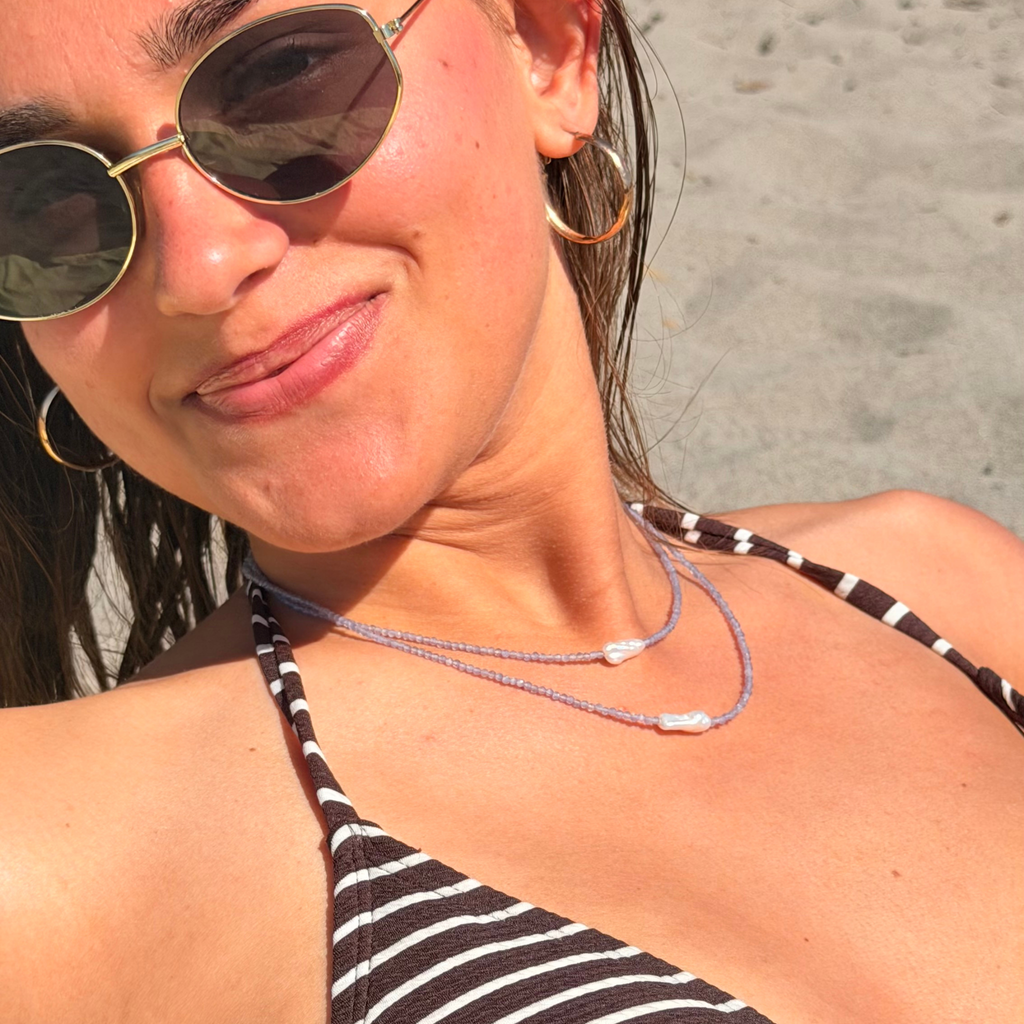 Woman wearing sunglasses and a striped bikini top on a beach and a pearl beaded necklace
